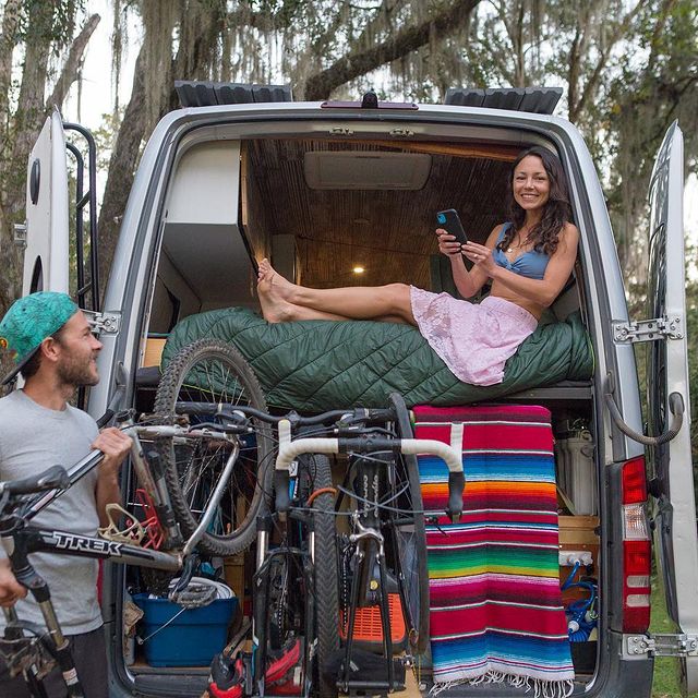 A smiling lady sitting on a permanent raised platform bed and a smiling man putting bikes into a camper van under bed garage