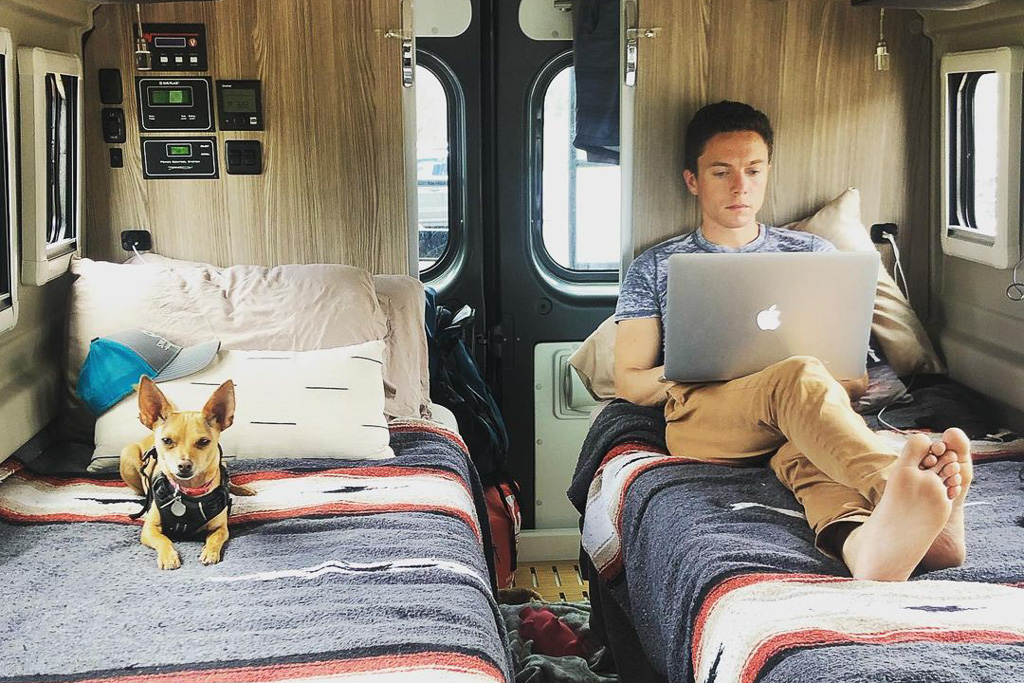 A man working on an apple laptop, sitting on a single bed in a camper van conversion.