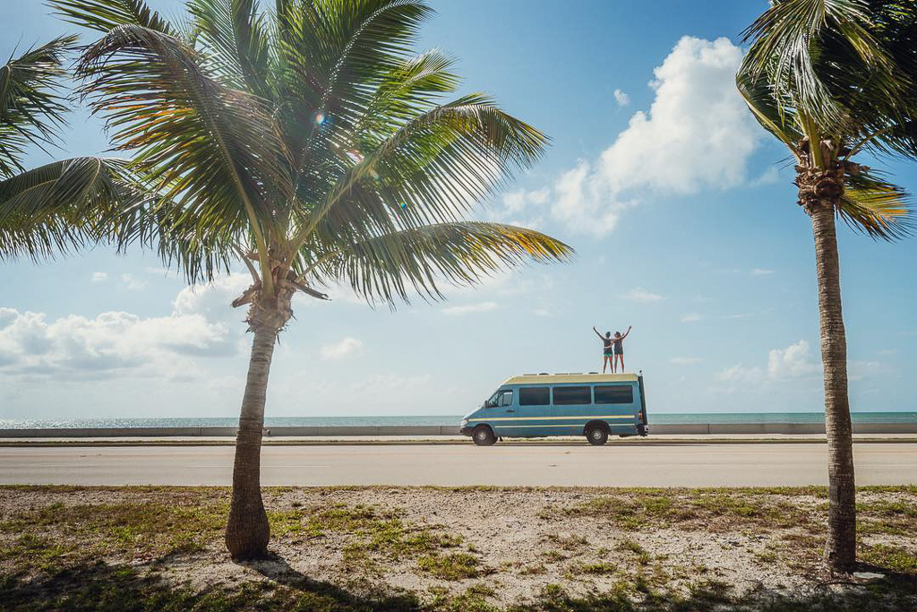 A couple standing on top of their campervan on a sunny beach with palm trees along it.