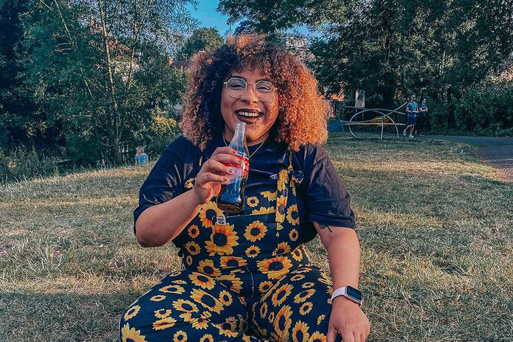 A lady with sunflower dungarees drinking a bottle of coke