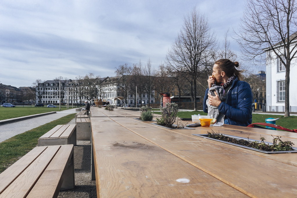A man biting an apple sitting at some long outdoor benches in Koblenz